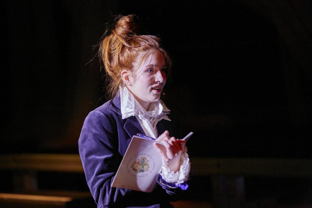 Person mit hochgestecktem Haar und dunklem Sakko hält ein Notizbuch und einen Stift in den Händen