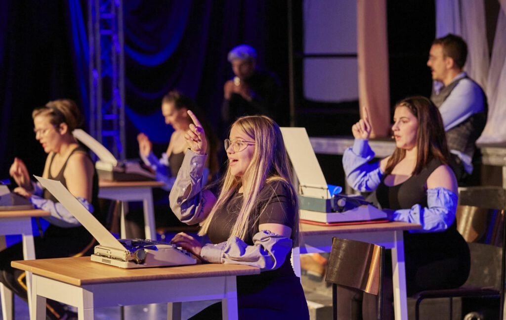 Lycantropia - Kein Weg zurück (2024) Mehrere Personen sitzen an Tischen mit Schreibmaschinen und heben jeweils eine Hand mit ausgestrecktem Zeigefinger