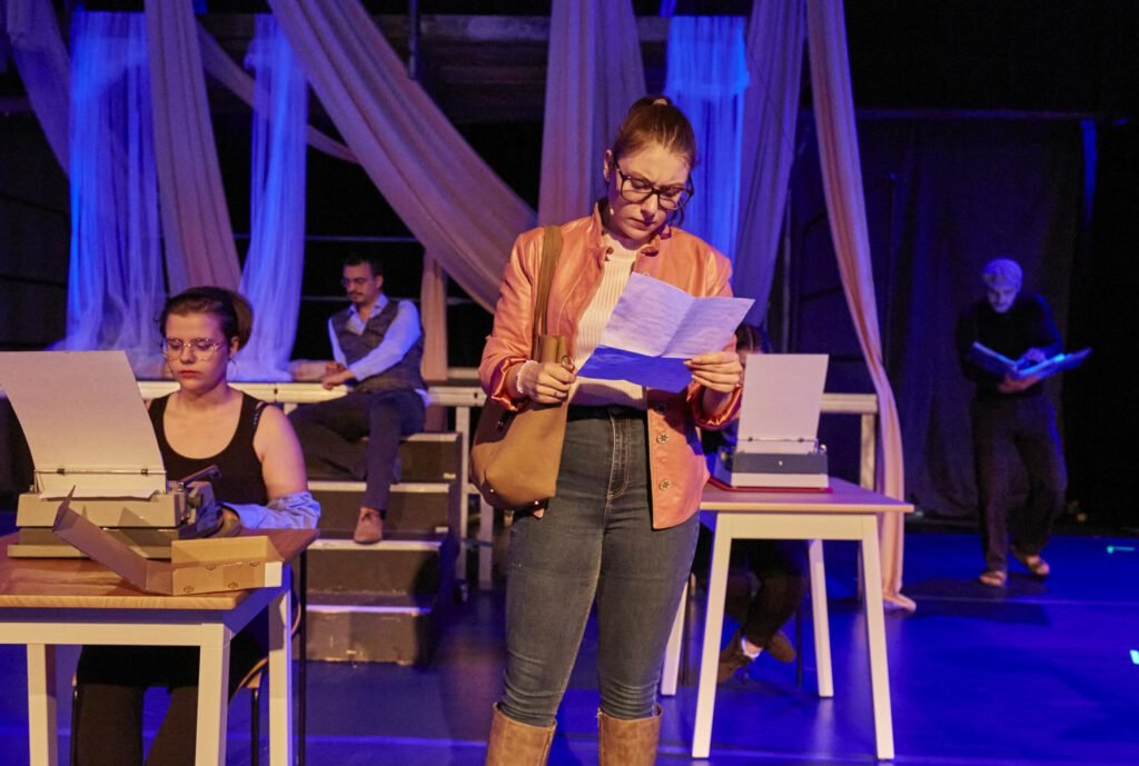 Frau steht auf einer Bühne und liest ein Blatt Papier vor, im Hintergrund sitzen und stehen weitere Personen mit Schreibmaschinen und Büchern