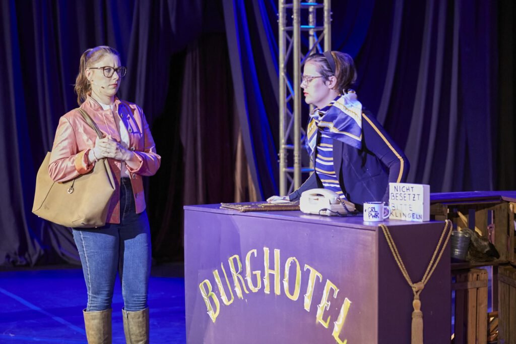 Zwei Frauen in einem Bühnenbild mit einem Tresen, auf dem "BURGHOTEL" steht, eine Frau steht mit Tasche, die andere lehnt am Tresen mit Telefon