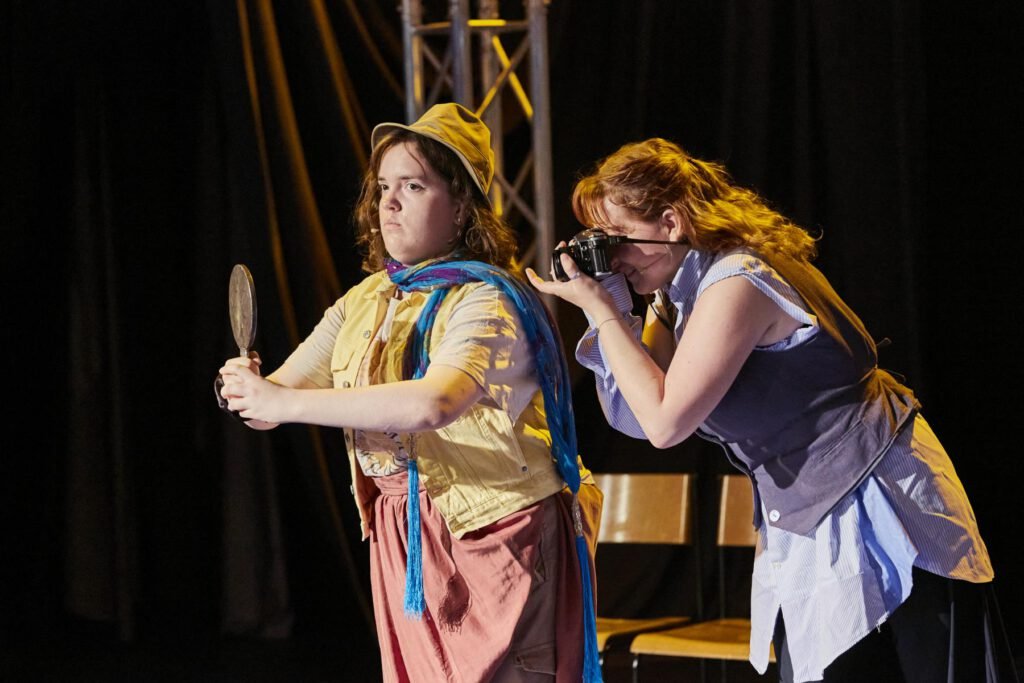 Lycantropia - Kein Weg zurück (2024) Zwei Frauen auf einer Bühne, eine hält einen Handspiegel, die andere fotografiert mit einer Kamera, gelbes Licht