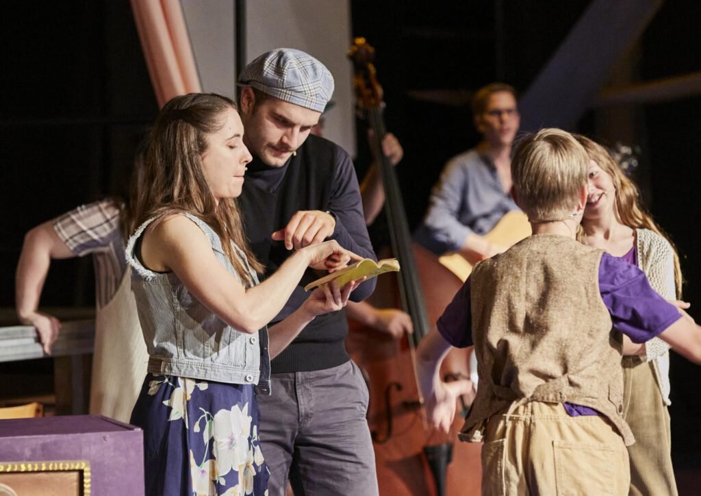 Lycantropia - Kein Weg zurück (2024) Mehrere Personen stehen auf einer Bühne, eine Frau hält ein Buch, zwei Kinder tanzen, im Hintergrund ist ein Kontrabass zu sehen.