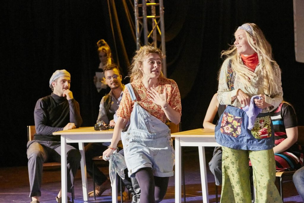 Lycantropia - Kein Weg zurück (2024) Mehrere Personen sitzen und stehen auf einer Bühne, im Café, zwei Frauen im Vordergrund tragen bunte Kleidung und bewegen sich.