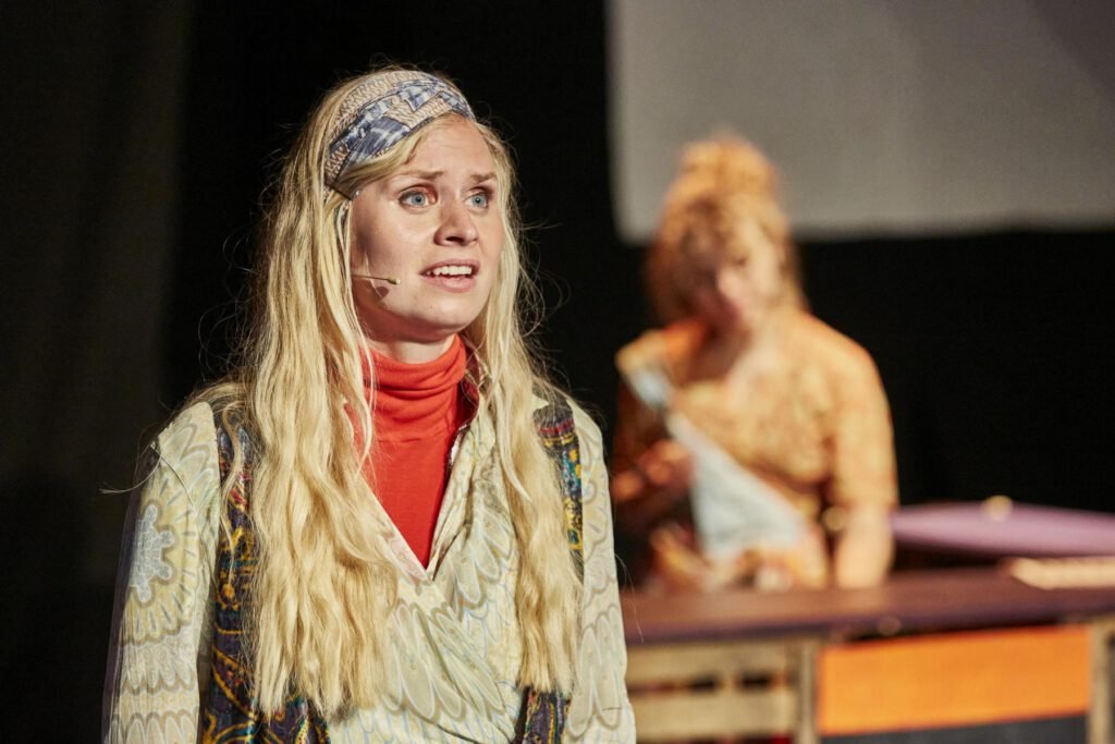 Lycantropia - Kein Weg zurück (2024) Frau mit langem, gewelltem Haar und einem Oberteil mit Blumenmustern steht im Vordergrund und singt, unscharfe Person im Hintergrund steht hinter einem Cafe-Tresen