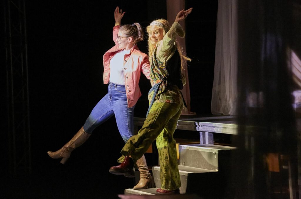 Zwei Frauen stehen einer Treppe auf einer Bühne, beide heben ein Bein und jeweils einen Arm nach oben. Sie tanzen.