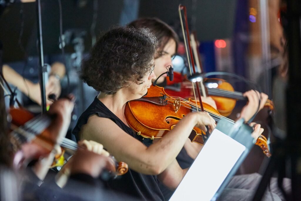 Mehrere Musiker spielen Geige in einem Orchester, eine Frau im Vordergrund mit lockigem Haar und schwarzem Oberteil hält eine Geige und den Bogen
