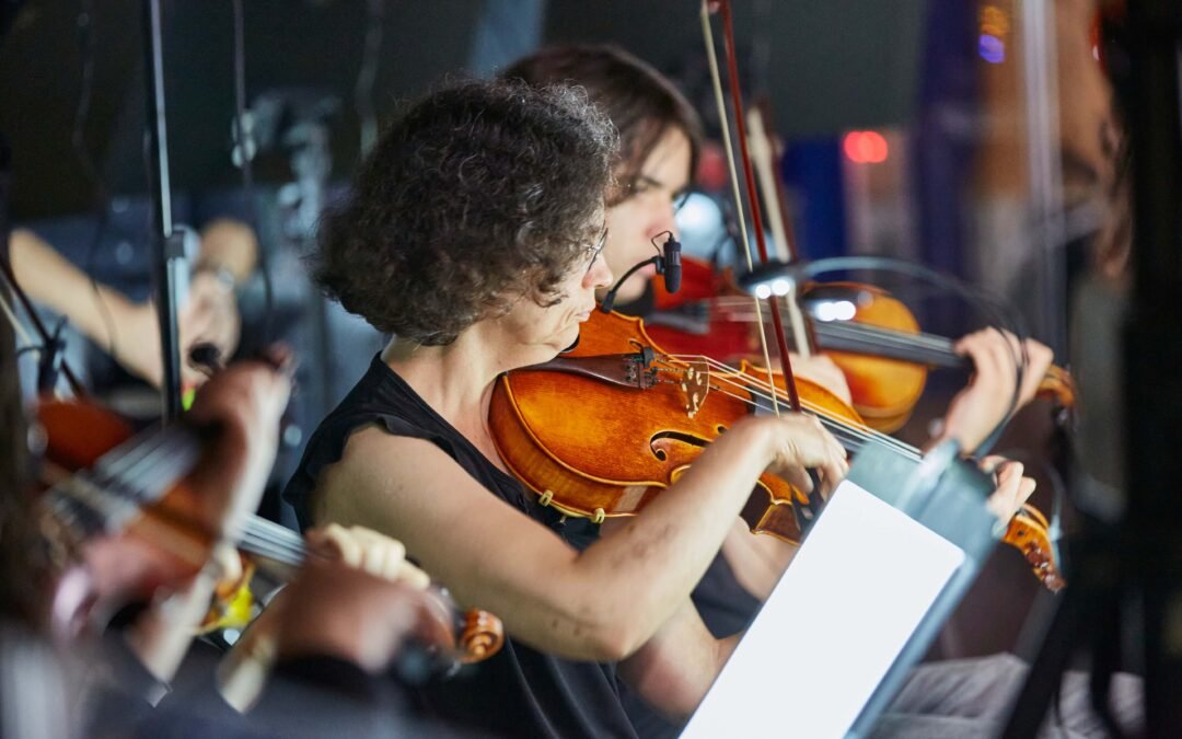 Backstage-1 Mehrere Musiker spielen Geige in einem Orchester, eine Frau im Vordergrund mit lockigem Haar und schwarzem Oberteil hält eine Geige und den Bogen