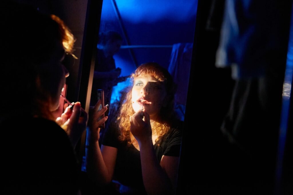 Person mit lockigem Haar trägt vor einem Spiegel Lippenstift auf