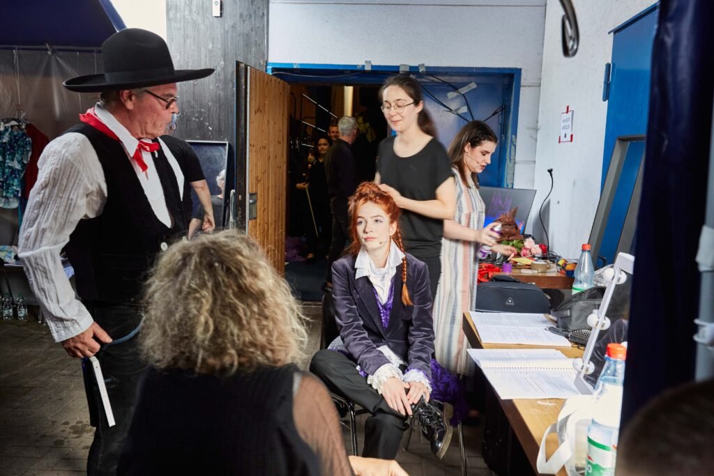 Mehrere Personen mit Theaterkostümen und Alltagskleidung in einem Backstagebereich mit Spiegeln, Tischen und Requisiten