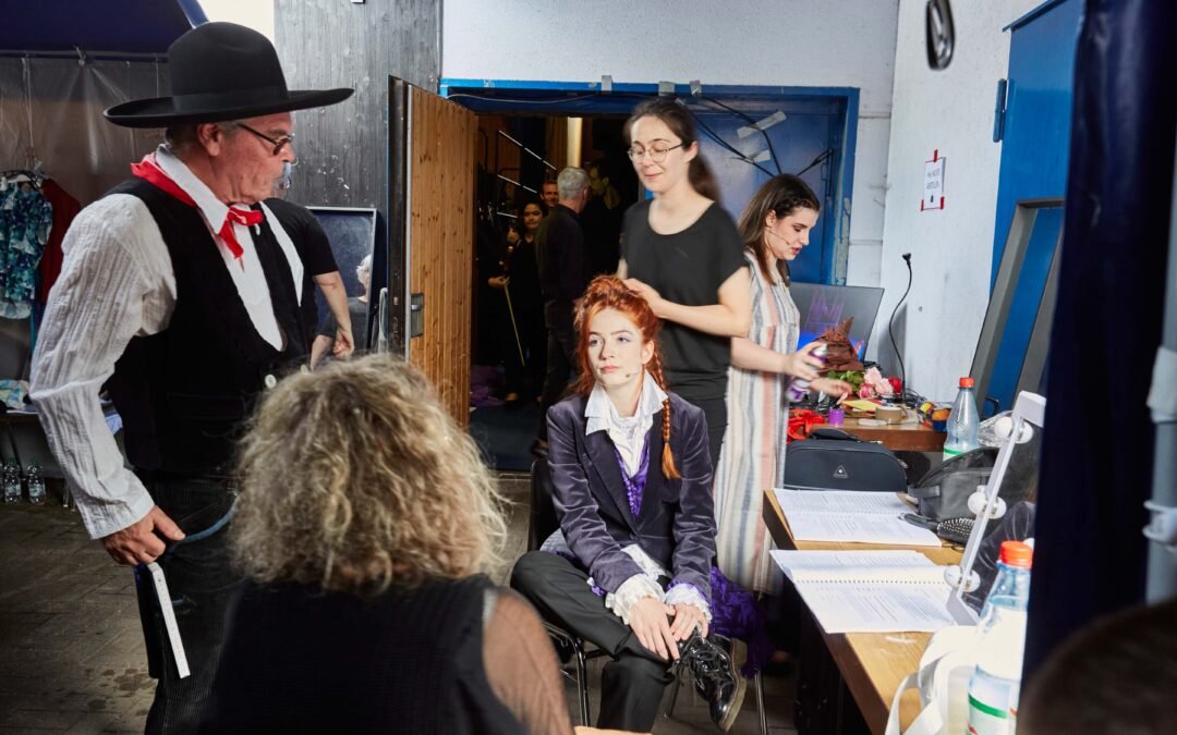 Backstage-2 Mehrere Personen mit Theaterkostümen und Alltagskleidung in einem Backstagebereich mit Spiegeln, Tischen und Requisiten