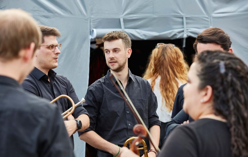 Gruppe von Musikern in schwarzer Kleidung mit Blechblasinstrumenten steht vor einem Zelt und unterhalten sich