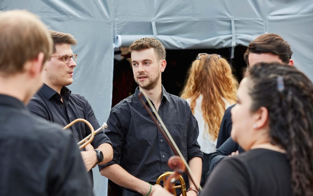 Backstage-3 Gruppe von Musikern in schwarzer Kleidung mit Blechblasinstrumenten steht vor einem Zelt und unterhalten sich