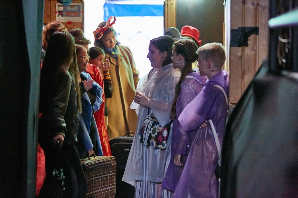 Gruppe von Menschen in bunten Kostümen steht dicht gedrängt in einem schmalen Flur mit Koffern und Taschen