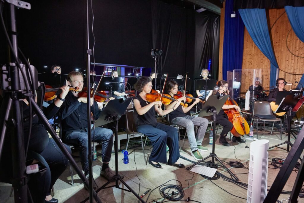 Backstage-6 Gruppe von Musikerinnen und Musikern sitzt mit Streichinstrumenten auf Stühlen vor Notenständern in einem Raum mit Holzverkleidung und blauen Vorhängen