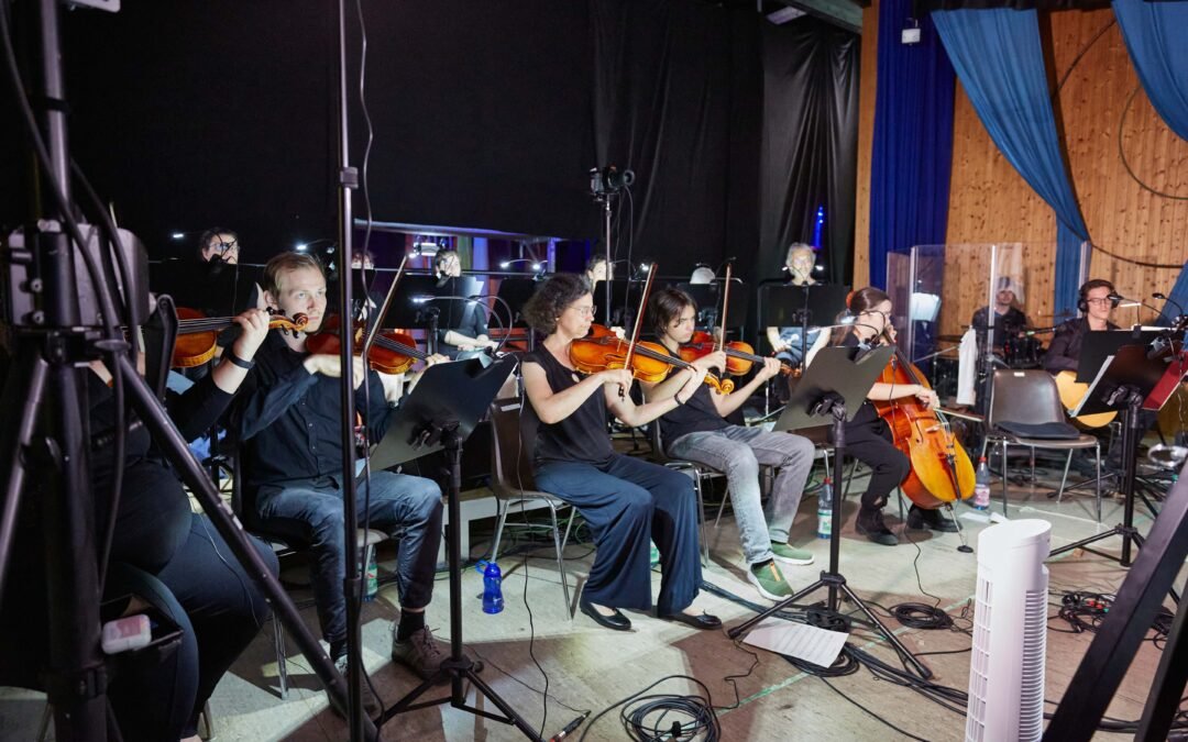 Backstage-6 Gruppe von Musikerinnen und Musikern sitzt mit Streichinstrumenten auf Stühlen vor Notenständern in einem Raum mit Holzverkleidung und blauen Vorhängen