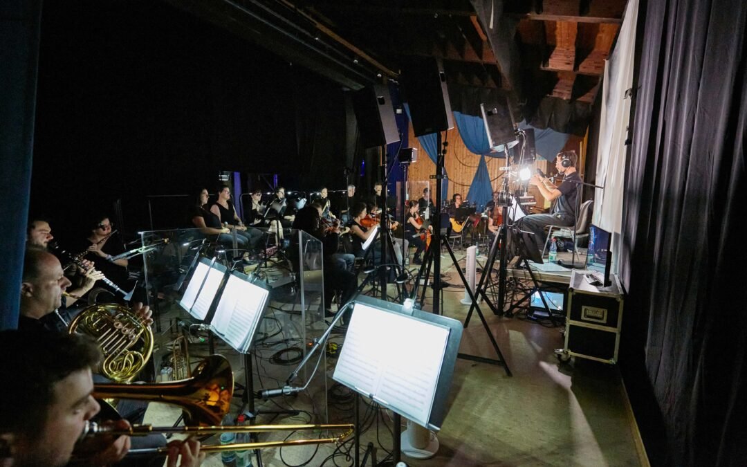 Backstage-7 Orchester mit verschiedenen Instrumenten und einem Dirigenten, der mit Kopfhörern vor einem Mikrofon sitzt