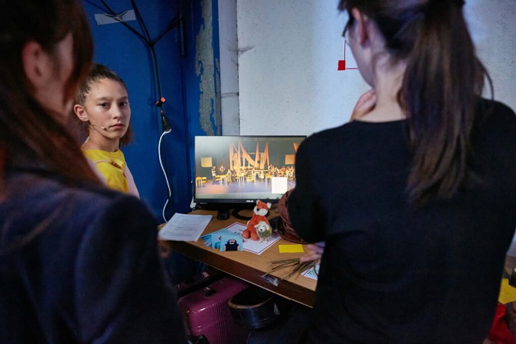 Drei Personen von hinten betrachten einen Monitor, der das Geschehen auf der Bühne zeigt