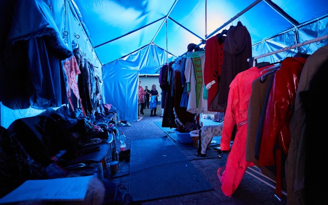 Backstage-9 Blick in einen Zeltgang mit an beiden Seiten hängenden Kostümen und zwei Personen im Hintergrund