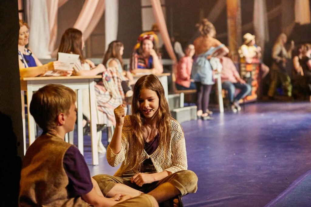 Kinder sitzen mit verschränkten Beinen auf dem Boden und spielen miteinander, während andere Personen im Hintergrund an Tischen sitzen oder stehen