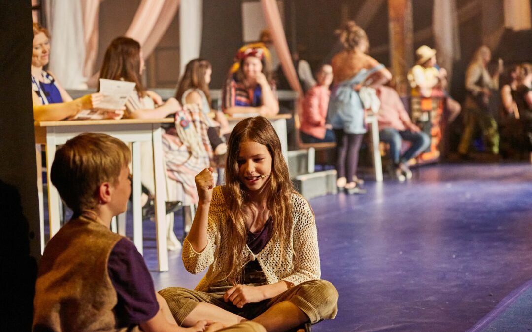 Kinder sitzen mit verschränkten Beinen auf dem Boden und spielen miteinander, während andere Personen im Hintergrund an Tischen sitzen oder stehen