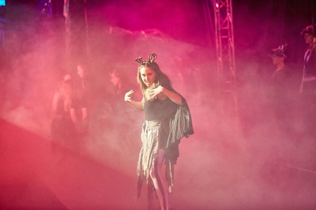 Lycantropia-08 Person mit Kopfschmuck und zerrissenem Rock steht in rotem Bühnennebel vor mehreren im Hintergrund stehenden Personen