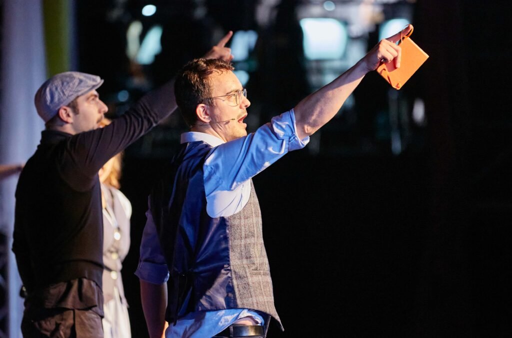 Lycantropia-05 Zwei Männer stehen nebeneinander, sie heben beide einen Arm mit ausgestrecktem Zeigefinger nach oben, eine Person hält ein oranges Notizbuch in der Hand