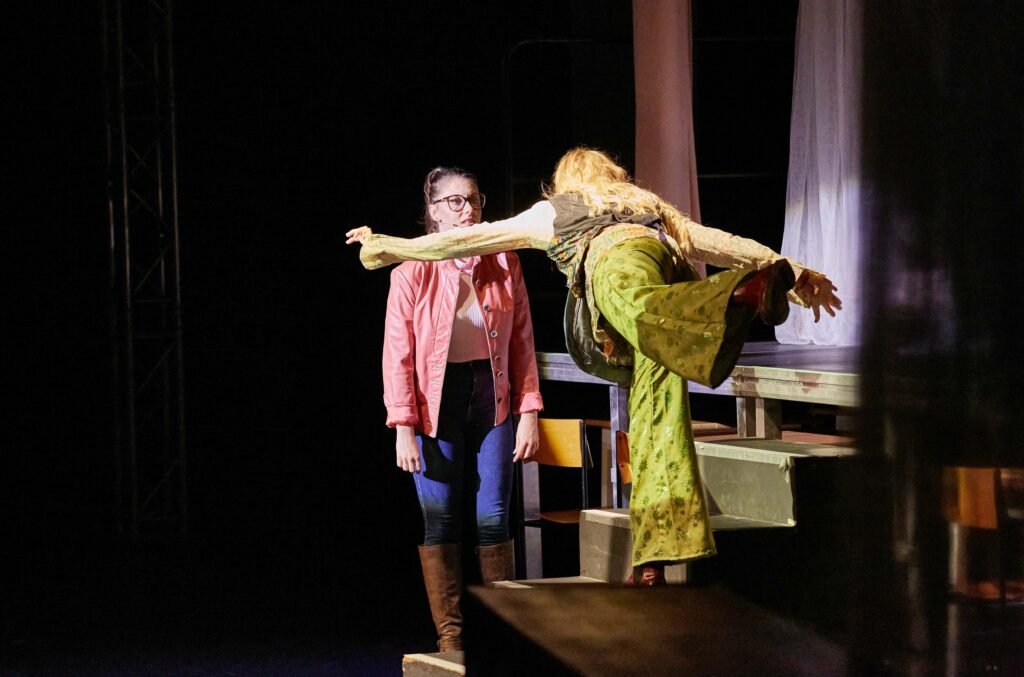 Lycantropia-03 Eine Frau in grüner Hose und langem Haar balanciert mit ausgestrecktem Bein auf einer Bühne vor einer stehenden Person in rosa Jacke und Stiefeln