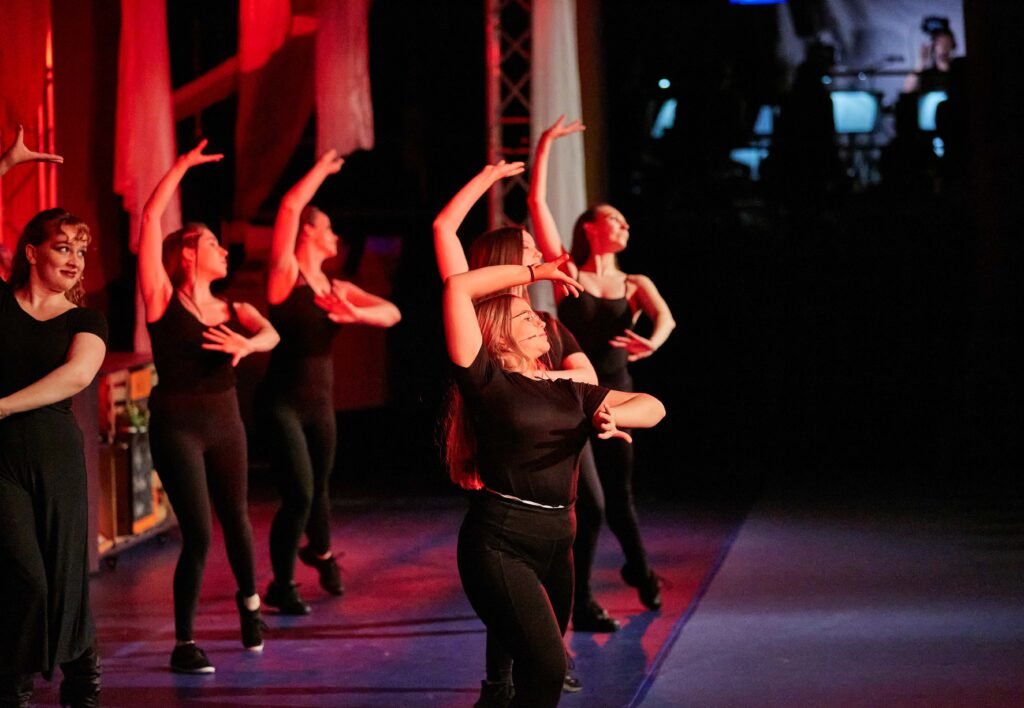 Gruppe von Tänzerinnen in schwarzer Kleidung mit erhobenen Armen auf einer Bühne mit roter Beleuchtung